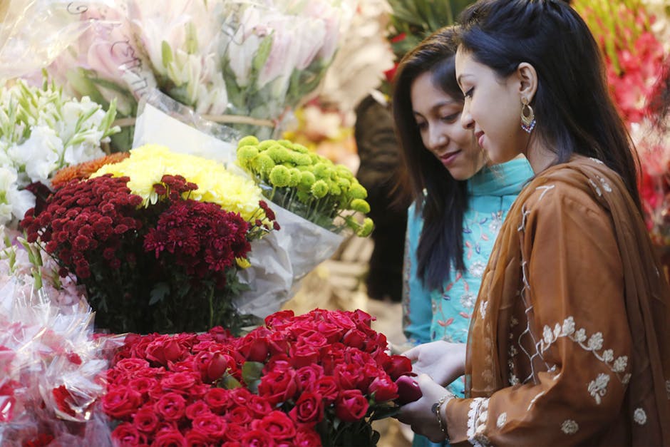 Two women are shopping for flowers at a florist, with a vibrant selection of fresh floral bouquets and arrangements displayed around them. One woman, wearing a brown traditional dress with white embro