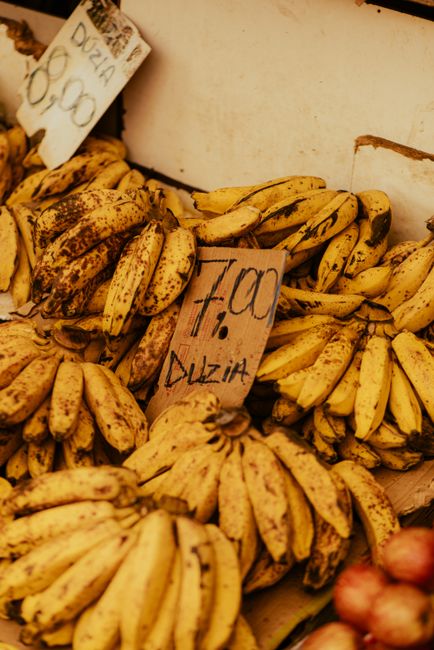 A close-up image of ripe bananas displayed for sale, with a yellow peel marked by brown speckles indicating ripeness, arranged in bunches and resting on a market stall surface. The bananas are placed