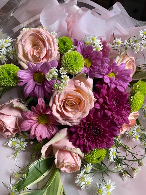 A close-up view of a floral bouquet arranged with large, pale pink roses featuring soft, layered petals, complemented by vibrant purple daisies with yellow centers, bright green button chrysanthemums,